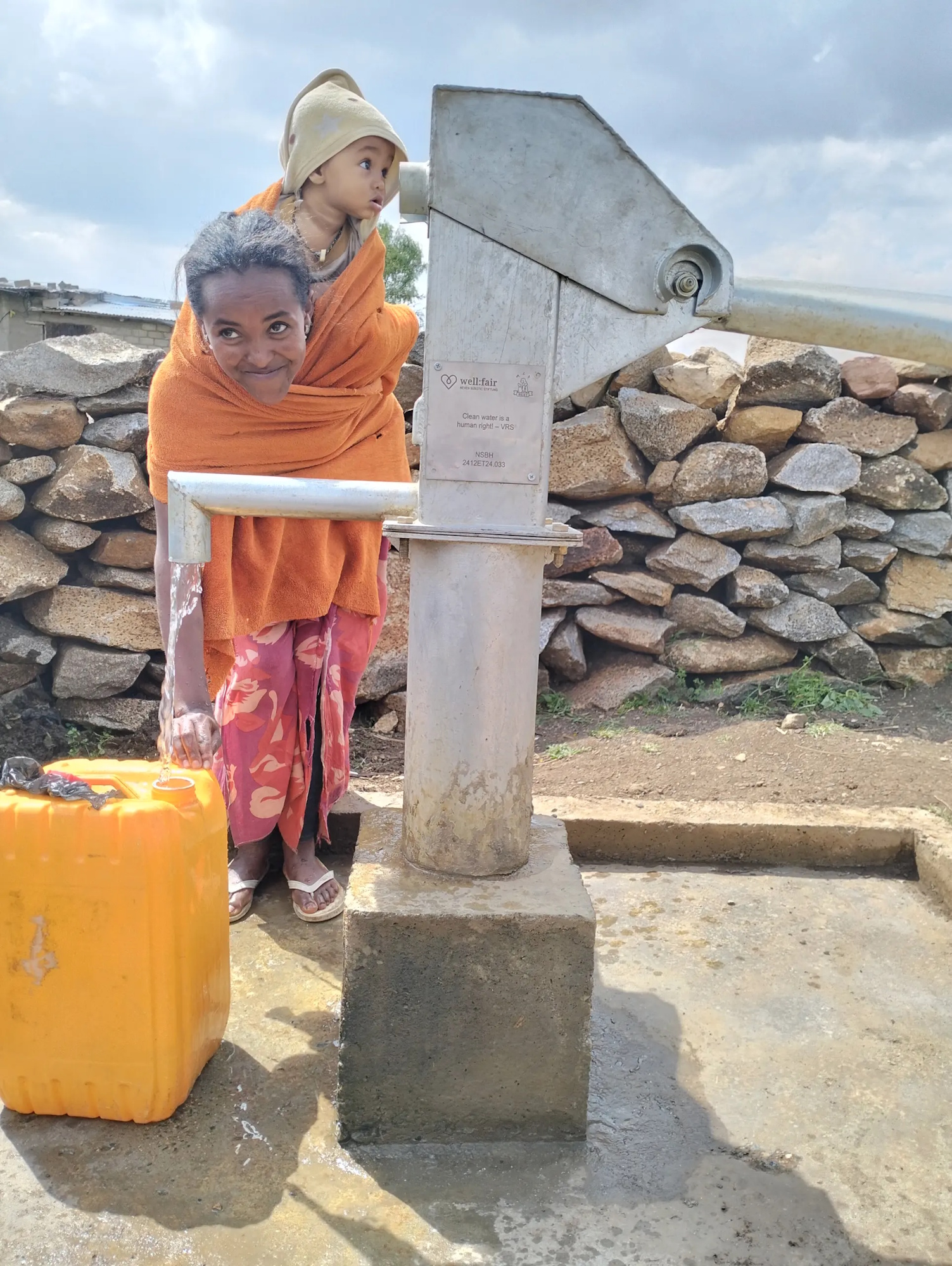 Das Bild zeigt eine Mutter mit Kind, die sich über das saubere Trinkwasser aus dem neugebauten Brunnen freuen (© well:fair foundation)