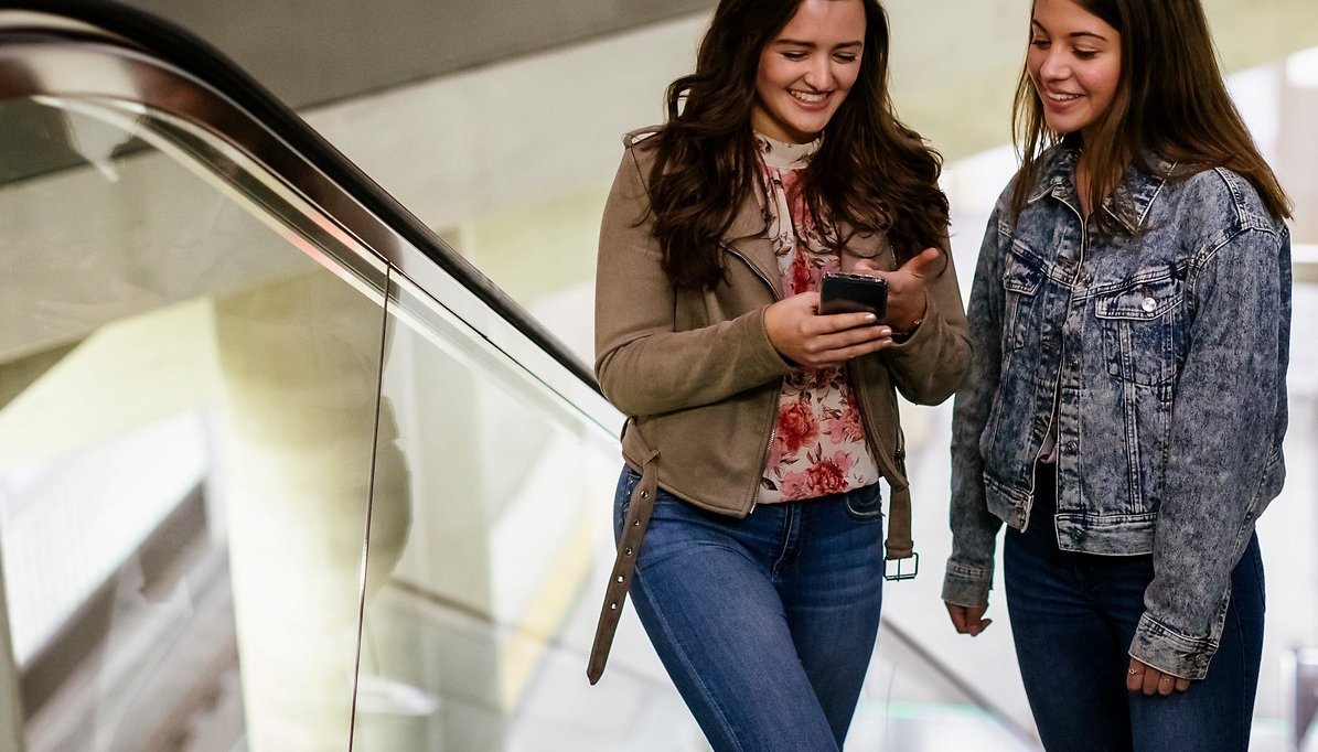 Das Bild zeigt zwei junge Frauen mit Handy in der Hand auf der Rolltreppe, Haltestelle Köln Chlodwigplatz