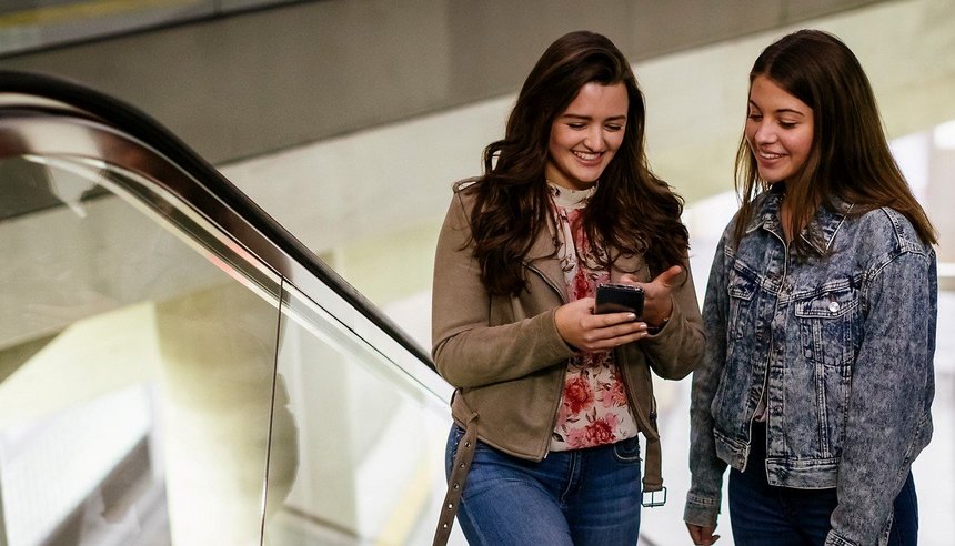 Das Bild zeigt zwei junge Frauen mit Handy in der Hand auf der Rolltreppe, Haltestelle Köln Chlodwigplatz